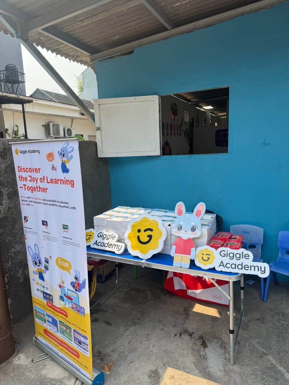 A folding table outside a school with a bright blue wall, a Giggle Academy roll-up banner, stacked boxes, Giggle Academy signs and a cut-out of Tom the rabbit.