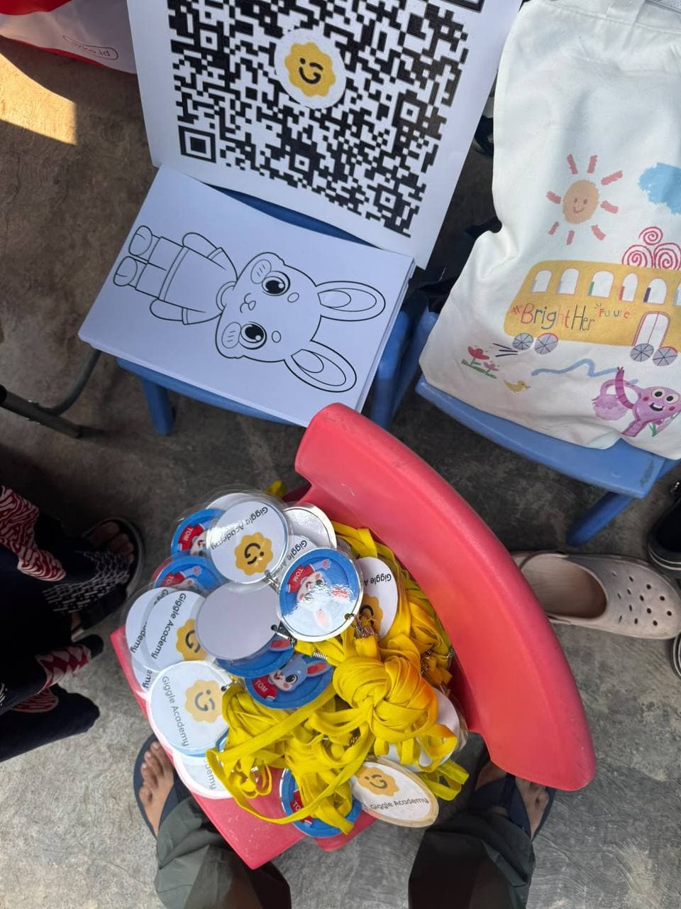 Seen from above: a small red chair piled with yellow lanyards carrying round Giggle Academy and TOM badges, a stack of Tom colouring sheets and a printed QR code on a blue chair behind it.
